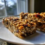 Close-up of chewy homemade granola bars packed with oats, seeds, and dried cranberries, sitting on a white plate.