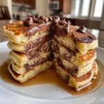 A stack of fluffy chocolate chip pancakes cut in half, showing melted chocolate filling and topped with chocolate chips and syrup.