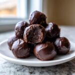 A stack of rich, chocolate-covered Oreo balls on a white plate, with one showing a bite taken out.