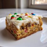 A thick, square slice of rich christmas cookie bars topped with white frosting and colorful M&M candies.
