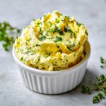 A close-up of fluffy, yellow cowboy butter dipping sauce mixed with green herbs in a white ramekin.