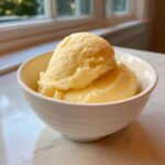 Two scoops of pale yellow banana ice cream served in a white bowl on a marble countertop.
