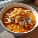 A close-up of a white bowl filled with creamy chicken tortilla soup, topped with shredded chicken, corn, cotija cheese, and crispy tortilla strips.