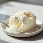 A close-up of a creamy scoop of white cottage cheese ice cream melting slightly on a white plate with a spoon taking a bite.