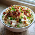 A white bowl filled with creamy Million Dollar Dip, generously topped with crumbled bacon and fresh green onions.