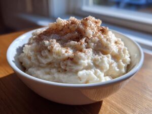 A close-up of a creamy bowl of rice pudding recipe, generously sprinkled with ground cinnamon, sitting on a wooden surface.