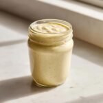 A glass jar filled with thick, creamy, yellowish homemade salad dressing, sitting on a marble surface.