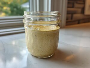 A creamy, light brown homemade salad dressing stored in a clear mason jar on a white marble counter.