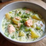 A close-up of a bowl of creamy salmon soup featuring chunks of pink salmon, potatoes, celery, and fresh dill.