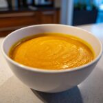 Close-up of a bowl filled with vibrant, creamy orange squash soup resting on a light countertop.