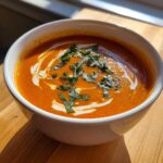 A close-up of rich, orange tomato basil soup swirled with cream and topped with fresh, chopped basil.