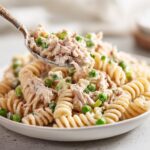 A spoonful of creamy tuna pasta salad with peas and celery being lifted from a white bowl.