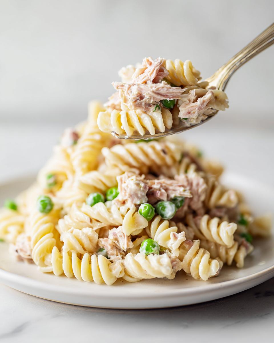 A spoonful of creamy tuna pasta salad with rotini pasta and green peas being lifted from a plate.