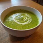Close-up of a bowl of vibrant green creamy zucchini soup topped with a white cream swirl.