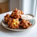 Four golden brown, crispy corn fritters served on a plate next to a small bowl of creamy dipping sauce.