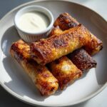 A pile of golden brown, crispy peach cobbler egg rolls served on a white plate next to a small ramekin of white dipping sauce.