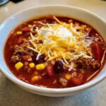 Close-up of a bowl of rich crockpot cowboy soup topped with shredded cheese, sour cream, corn, and beans.