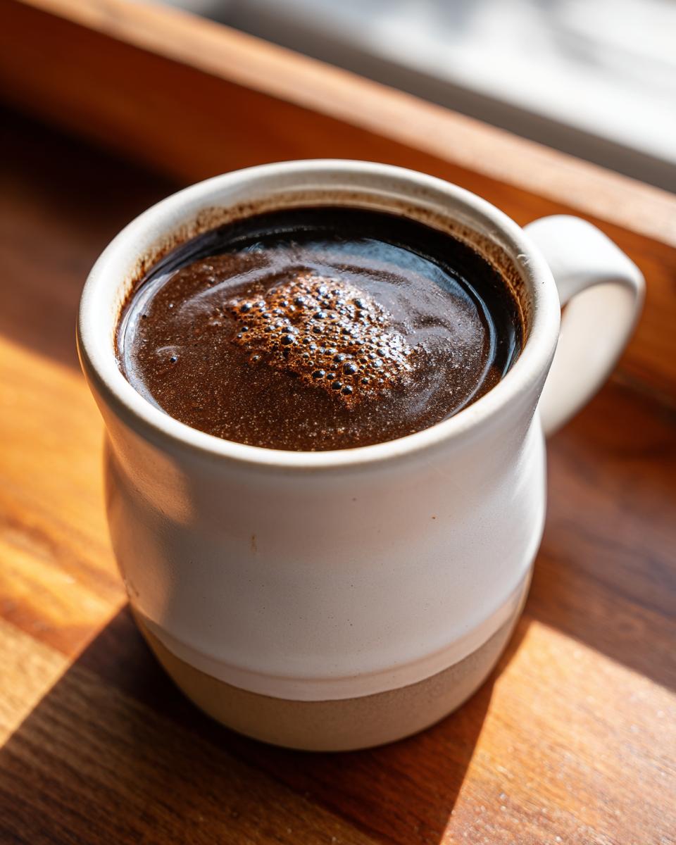 A dark, rich beverage, possibly coffee or a dark broth, in a white and tan ceramic mug next to a window, unrelated to black bean soup.