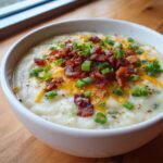 A close-up of a bowl of creamy easy crockpot potato soup topped generously with shredded cheddar cheese, crispy bacon bits, and fresh green onions.