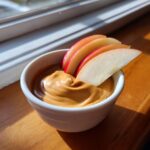 A small white bowl filled with creamy peanut butter fruit dip, garnished with fresh red apple slices.