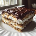 Close-up of a layered slice of eclair cake featuring graham crackers, whipped cream, and chocolate frosting.