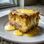 A square slice of rich eggs benedict casserole topped with dripping yellow hollandaise sauce on a white plate.