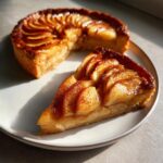 A close-up of a slice of flaky apple tart dusted with cinnamon, with the rest of the apple tart visible behind it.