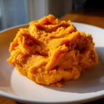 A close-up, appetizing serving of fluffy mashed sweet potatoes on a white plate, highlighted by sunlight.