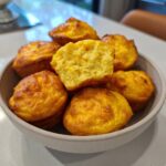A close-up of several golden brown, fluffy egg bites piled high in a light gray bowl, one cut in half showing the texture.