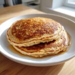 Two fluffy oatmeal pancakes stacked on a white plate, drizzled with maple syrup.