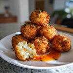 A stack of golden brown, crispy fried goat cheese balls, one broken open showing the melted interior, served with sweet chili sauce.