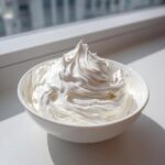 A white bowl filled with stiff, glossy, perfectly piped gingerbread house icing sitting on a white surface near a window.