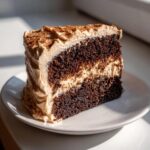 Close-up of a rich, dark slice of gingerbread latte cake layered with light brown frosting and dusted with spice.