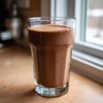 A tall glass filled with rich, frothy chocolate milk sitting on a light countertop near a window.