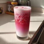 A tall glass of layered strawberry milk showing a gradient from white milk at the bottom to vibrant pink foam at the top.