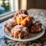 A stack of golden brown baked apple fritters generously drizzled with white vanilla glaze, sitting on a white plate.
