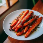 A close-up of glossy, glazed roasted carrots topped with herbs and coarse salt on a white plate.