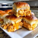 Close-up of gooey, cheesy buffalo chicken sliders stacked on a white platter, showing shredded chicken filling.