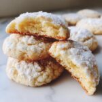A stack of freshly baked, soft gooey butter cookies dusted heavily with powdered sugar, one cookie is broken open showing the texture.
