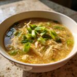 A close-up shot of a bowl of homemade egg drop soup, featuring wispy egg ribbons and topped with bright green scallions.