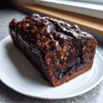 A rich, dark chocolate loaf of hot fudge brownie bread drizzled with melted chocolate, served on a white plate.