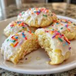 Close-up of soft Italian ricotta cookies with white glaze and colorful sprinkles, one cookie broken open.