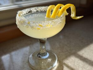 A bright yellow lemon drop cocktail in a sugar-rimmed coupe glass, garnished with a long lemon peel spiral.