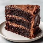 Close-up of a moist, three-layer slice of rich chocolate cake recipe with thick chocolate frosting.
