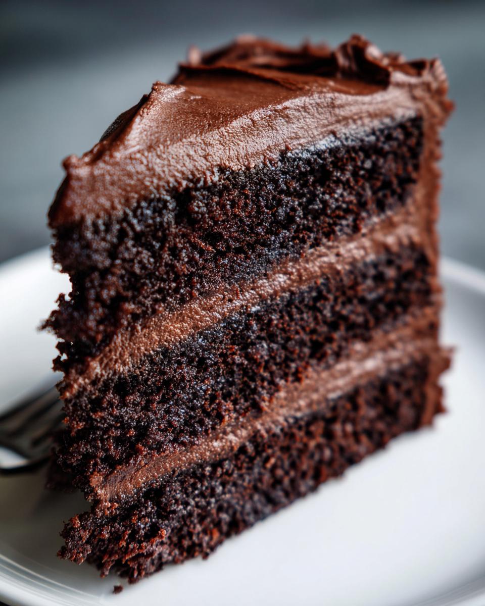 Close-up of a moist, three-layer slice of chocolate cake recipe covered in rich chocolate frosting.