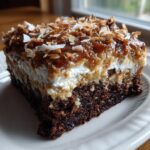 A close-up of a moist slice of german chocolate poke cake showing the dark chocolate base, white filling, and pecan-coconut topping.