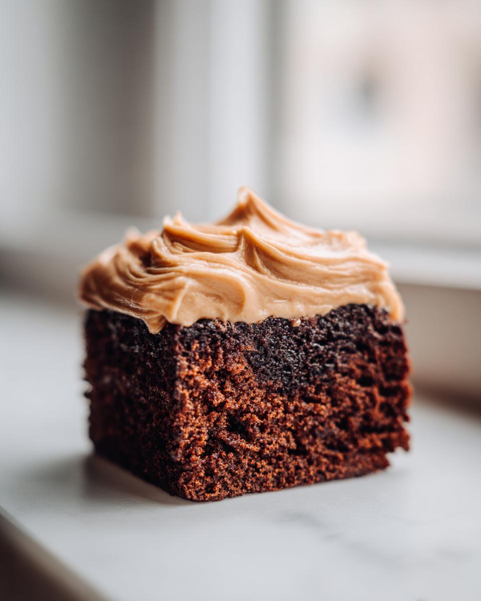 Close-up of a square slice of moist, dark gingerbread cake topped with swirled caramel-colored frosting.