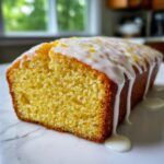 A close-up of a moist slice of lemon loaf cake topped with white citrus glaze and zest.