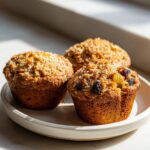 Three freshly baked morning glory muffins topped with coarse sugar and visible raisins, sitting on a light plate.