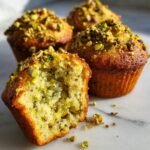 Close-up of a torn pistachio muffin showing the moist interior, topped with chopped pistachios.